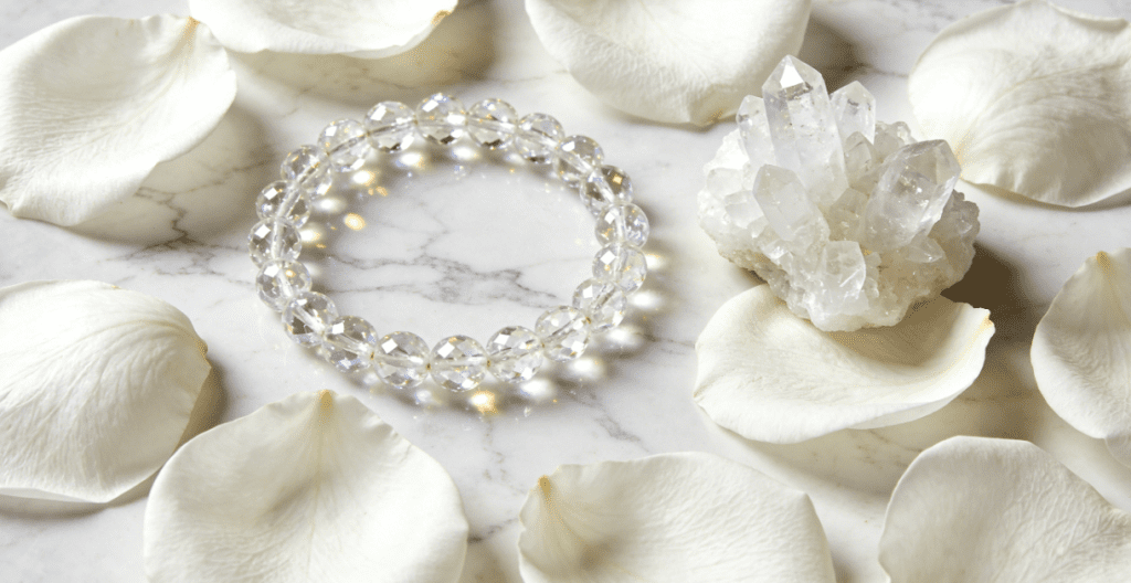 Crystal mala beads placed on marble countertop covered with fresh white rose petals, accompanied by a clear quartz cluster